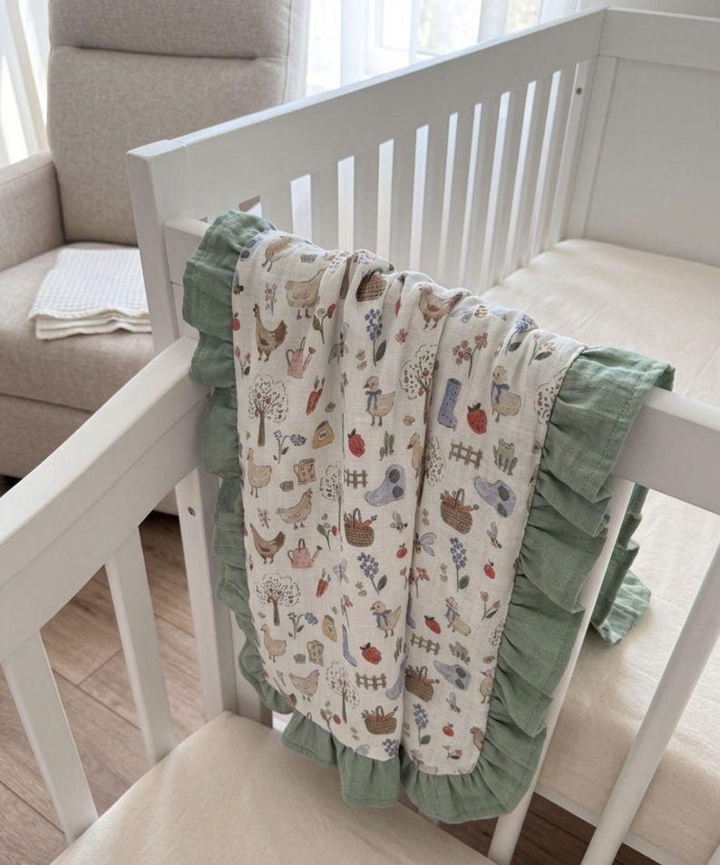 May include: A cream-colored baby blanket featuring a repeating pattern of chickens, ducks, trees, flowers, watering cans, and baskets. The blanket is finished with a ruffled sage green border along the edges and is draped over a white crib rail.