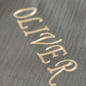 May include: A dark gray fabric with a crinkled texture featuring the name OLIVER embroidered in a light gold thread. The letters are stitched in a clean, sans-serif font, creating a subtle contrast against the dark, textured background material.