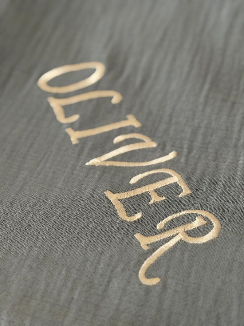 May include: A dark gray fabric with a crinkled texture featuring the name OLIVER embroidered in a light gold thread. The letters are stitched in a clean, sans-serif font, creating a subtle contrast against the dark, textured background material.