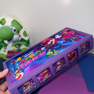 Puede incluir: Una caja rectangular de color morado con gr&aacute;ficos de Super Mario Galaxy en la tapa, incluyendo a Mario, Yoshi e im&aacute;genes espaciales. El lateral de la caja contiene cuatro ventanas cuadradas llenas de peque&ntilde;as cuentas de colores.