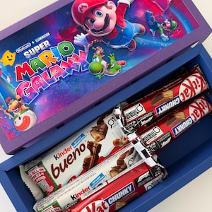 Puede incluir: Una caja de regalo azul con un gr&aacute;fico de Super Mario Galaxy en la tapa, que contiene dos barras de chocolate KitKat Chunky y dos barras Kinder Bueno en su interior. La tapa muestra a los personajes Mario, Luigi, Peach y Yoshi.