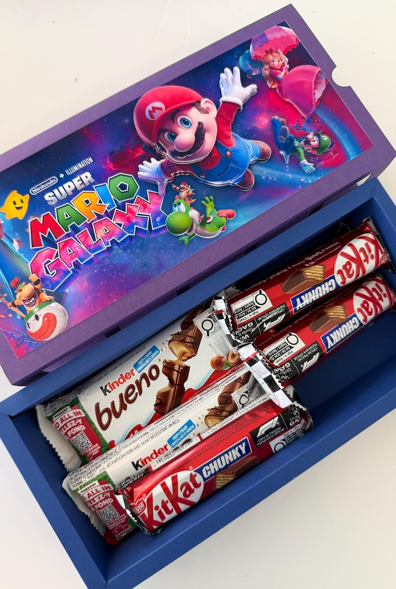 Puede incluir: Una caja de regalo azul con un gr&aacute;fico de Super Mario Galaxy en la tapa, que contiene dos barras de chocolate KitKat Chunky y dos barras Kinder Bueno en su interior. La tapa muestra a los personajes Mario, Luigi, Peach y Yoshi.