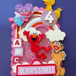 May include: A layered cake topper featuring Sesame Street characters Abby Cadabby, Big Bird, Elmo, and Zoe. It includes a rainbow, clouds, blocks with A, B, and C, a glittery number 4, and a pink street sign reading 123 OLIVIA'S STREET.