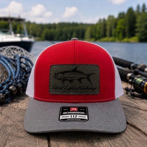 May include: A red, white, and gray trucker hat with a fish graphic and the text "Best Life Fishing" on a leather patch. The hat has a curved brim and an adjustable snap closure. The Richardson 112 trucker hat is displayed on a wooden surface.