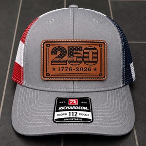 Könnte beinhalten: Eine graue Trucker-Cap mit einem rechteckigen braunen Lederaufnäher, der die Zahl 250 und den Text 1776-2026 zeigt. Die Kappe hat ein rot-weiß-blaues Netzteil und einen schwarzen Aufkleber auf dem Schirm mit der Aufschrift Richardson 112 Trucker.