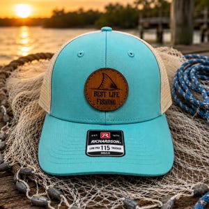 Puede incluir: Gorra de camionero turquesa y beige con un parche de cuero con una aleta de tiburón y el texto "BEST LIFE FISHING". La gorra tiene una visera curva y una parte trasera de malla. La etiqueta Richardson es visible.