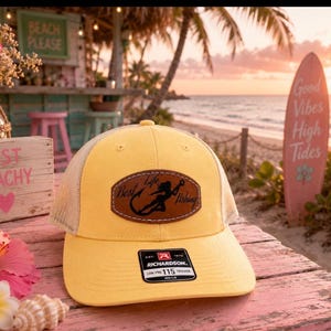 May include: A yellow trucker hat with a brown leather patch that reads "Best Life Fishing." The hat has a mesh back and a curved brim. The hat is sitting on a pink wooden surface with beach-themed decor in the background.
