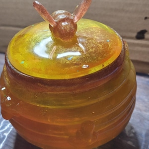 May include: A round, translucent yellow resin honey pot with a ribbed exterior and a lid featuring a sculpted bee topper. The material contains embedded gold glitter and small iridescent flakes, creating a shimmering effect throughout the container.