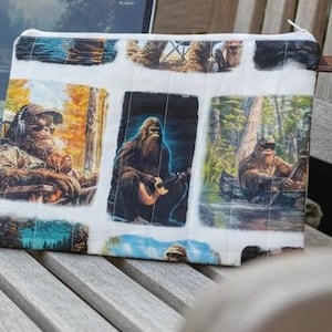 May include: A rectangular zipped pouch featuring a white background with a repeating grid of printed illustrations depicting a hairy creature in various outdoor settings, including playing a guitar, sitting in a canoe, and wearing headphones.
