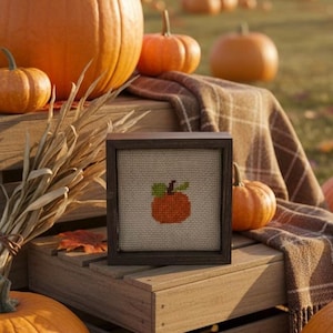 May include: A square wooden frame containing a cross-stitch embroidery of an orange pumpkin with a green leaf and brown stem on a beige fabric background. The frame is dark brown and features a simple, clean-lined design with a flat profile.