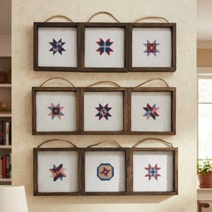 May include: A set of nine square wall art pieces arranged in three rows of three. Each piece features a dark brown wooden frame, a white fabric background with a colorful cross-stitched star pattern, and a jute twine loop for hanging.