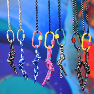 Peut inclure: Une collection de sept colliers composés de chaînes métalliques fixées à des mousquetons ovales de différentes couleurs, notamment jaune, bleu et rose. Chaque mousqueton retient un cordon en nylon à motifs noué par un simple nœud.