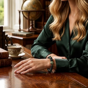 May include: A beaded bracelet featuring dark, textured volcanic stone beads and a central spherical charm with a metallic gold-toned cross design. The bracelet is worn on a wrist, showcasing the contrast between the matte black beads and the gold accent.