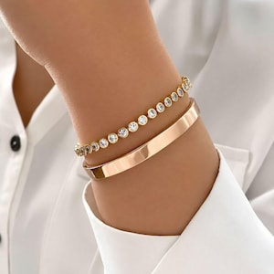 May include: Two gold bracelets are displayed. One is a plain, polished band. The other is a delicate tennis bracelet with small, round, clear stones. The bracelets are worn on a wrist, against the backdrop of a white shirt.