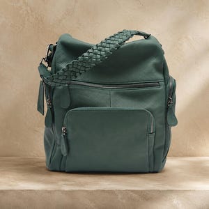 Op de afbeelding: Een blauwgroene leren rugzak met een gevlochten handvat, een groot voorvak met ritssluiting en extra zijvakken met ritssluiting. De tas is voorzien van zilverkleurige metalen hardware, ritstrekkers en een bijpassend leren label.