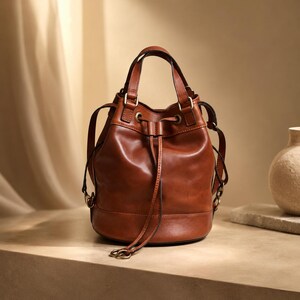 Pu&ograve; includere: Una borsa a secchiello in pelle marrone con chiusura a coulisse dotata di due lunghi tiranti in pelle, un manico superiore e attacchi laterali per una tracolla. La borsa presenta una base arrotondata, finiture in metallo ottonato e cuciture.