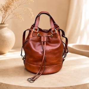 Pu&ograve; includere: Una borsa a secchiello in pelle color cognac con chiusura a coulisse, due lunghe stringhe in pelle, due manici superiori e minuteria metallica color ottone anticato. La borsa presenta una base circolare strutturata e attacchi laterali.