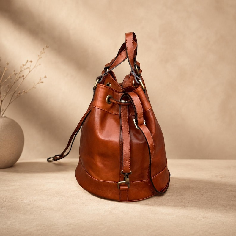 Pu&ograve; includere: Una borsa a secchiello in pelle color cognac con chiusura a coulisse, base circolare strutturata e minuteria in metallo color ottone anticato. La borsa include un manico superiore e una tracolla regolabile fissata con clip in metallo.