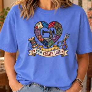 May include: A periwinkle blue t-shirt featuring a graphic of a heart-shaped quilt pattern containing a sewing machine, a spool of thread, and scissors. A yellow measuring tape wraps around the base with a banner reading SEW. CREATE. LOVE.