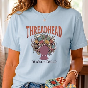 May include: A light blue t-shirt featuring a graphic of a silhouette head with hair made of tangled thread and colorful spools. The text reads THREADHEAD at the top and CREATIVELY TANGLED at the bottom in dark lettering. The design is centered on the chest.