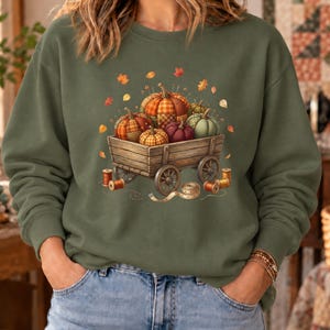 May include: An olive green crewneck sweatshirt featuring a graphic print of a wooden wagon filled with various patterned pumpkins, surrounded by scattered autumn leaves, wooden spools of thread, and a measuring tape, all centered on the chest.
