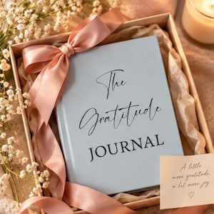 May include: A light blue hardcover journal with the text The Gratitude JOURNAL printed in black, nestled in a gift box with a dusty pink satin ribbon and a small card featuring the text A little more gratitude, a lot more joy.