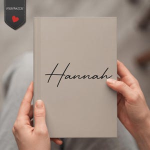 May include: A light beige hardcover notebook featuring the name Hannah written in a black cursive script font centered on the front cover. A small dark gray tag with the text PERSONALIZE and a red heart icon is positioned in the top left corner.