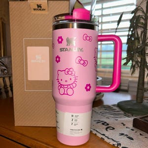 May include: A light pink Stanley tumbler with a dark pink Hello Kitty pattern featuring bows and flowers. The tumbler has a matching dark pink handle, a clear straw, and a white product label at the base. A cardboard Stanley box stands behind it.