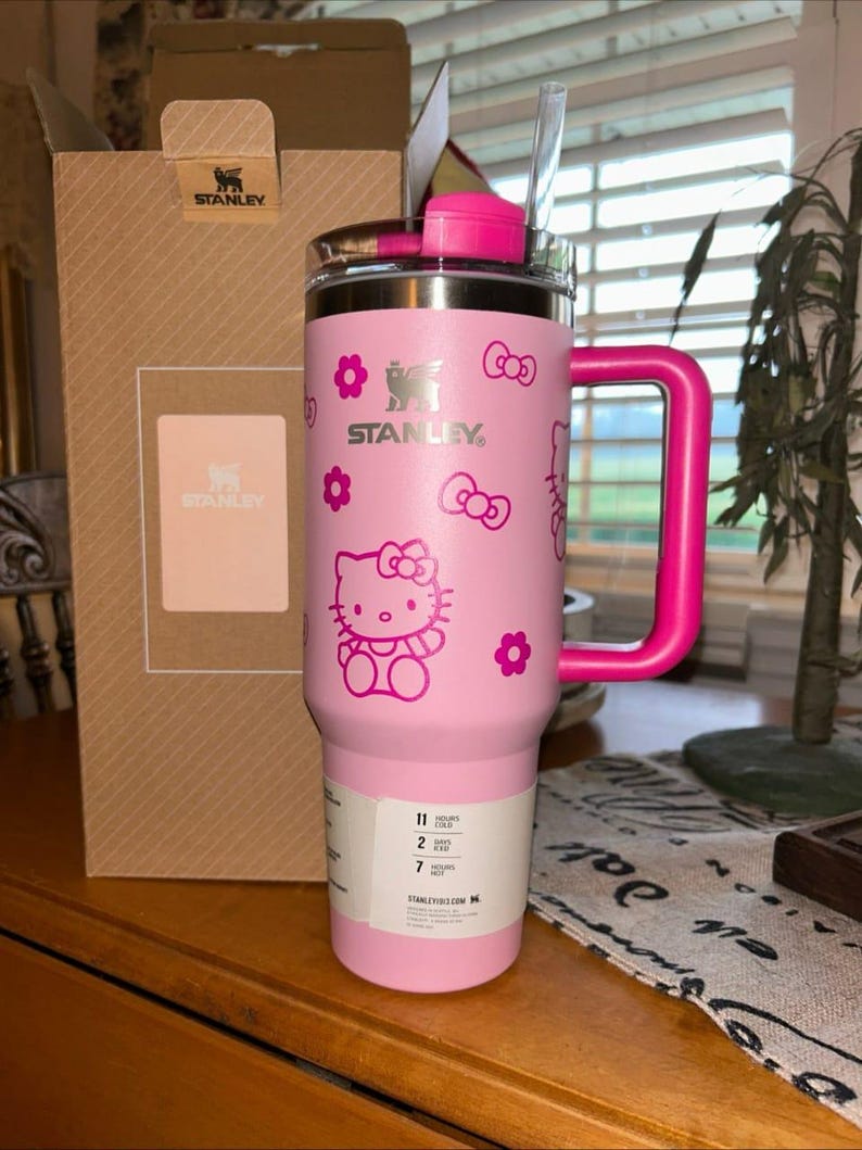 May include: A light pink Stanley tumbler with a dark pink Hello Kitty pattern featuring bows and flowers. The tumbler has a matching dark pink handle, a clear straw, and a white product label at the base. A cardboard Stanley box stands behind it.