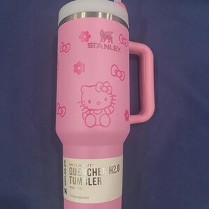 May include: A pink Stanley Quencher H2.0 tumbler with a 40 oz capacity, featuring a Hello Kitty graphic, flower icons, and a side handle. The tumbler includes a white lid with a straw and a product label that reads The FlowState Quencher H2.0.