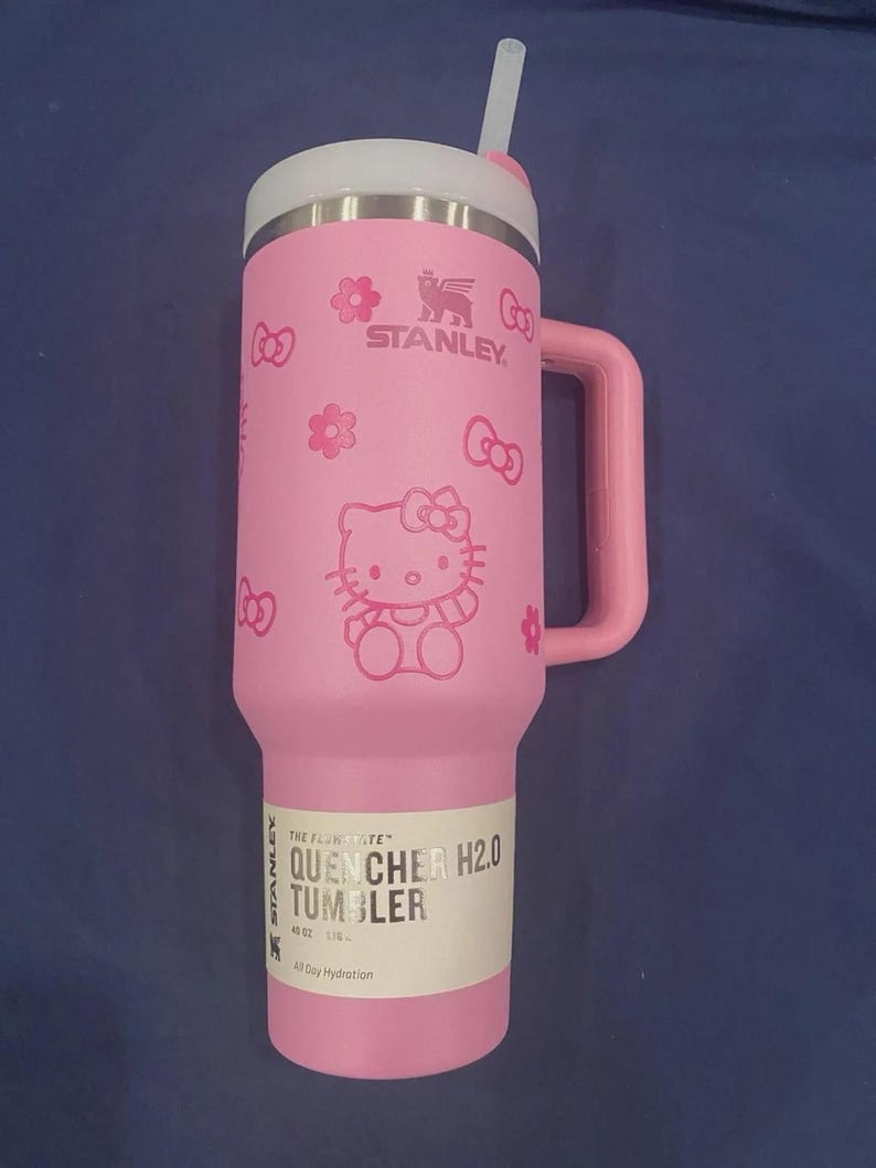 May include: A pink Stanley Quencher H2.0 tumbler with a 40 oz capacity, featuring a Hello Kitty graphic, flower icons, and a side handle. The tumbler includes a white lid with a straw and a product label that reads The FlowState Quencher H2.0.