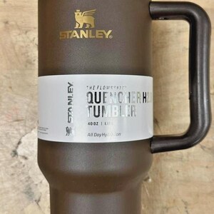 May include: Brown Stanley Quencher H2.0 Tumbler with a gold-colored rim and handle. The tumbler has the Stanley logo and the text "QUENCHER H2.0 TUMBLER" on a white label. It holds 40 oz (1.18 L) and is designed for all-day hydration.