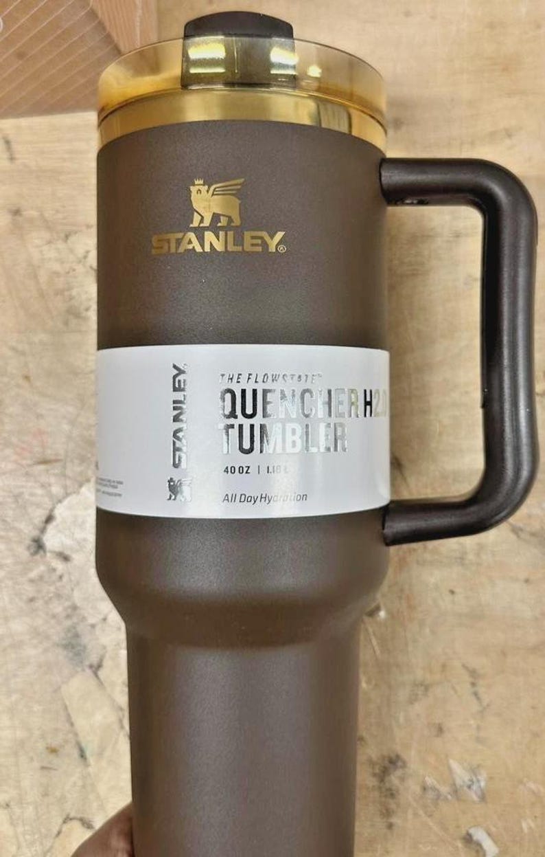 May include: Brown Stanley Quencher H2.0 Tumbler with a gold-colored rim and handle. The tumbler has the Stanley logo and the text "QUENCHER H2.0 TUMBLER" on a white label. It holds 40 oz (1.18 L) and is designed for all-day hydration.