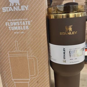 May include: A brown Stanley Flowstate tumbler with a gold rim and a straw, next to its cardboard box. The box features the Stanley logo and the text "The Quencher H2.0 Flowstate Tumbler 40 oz / 1.18 L". The tumbler is designed for all-day hydration.