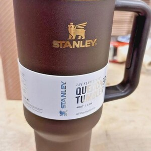 May include: Brown and gold Stanley Quencher Tumbler with a handle. The tumbler has the Stanley logo and the text "THE FLOWSTATE QUENCHER TUMBLER" on a white band. It holds 40 oz (1.18 L).