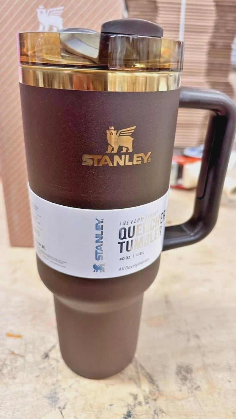 May include: Brown and gold Stanley Quencher Tumbler with a handle. The tumbler has the Stanley logo and the text "THE FLOWSTATE QUENCHER TUMBLER" on a white band. It holds 40 oz (1.18 L).