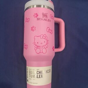 May include: A pink Stanley Quencher H2.0 tumbler featuring a Hello Kitty graphic with bows and flowers. The cup has a side handle, a white lid, and a clear straw. A product label on the lower section reads The FlowState Quencher H2.0 Tumbler 40 oz.