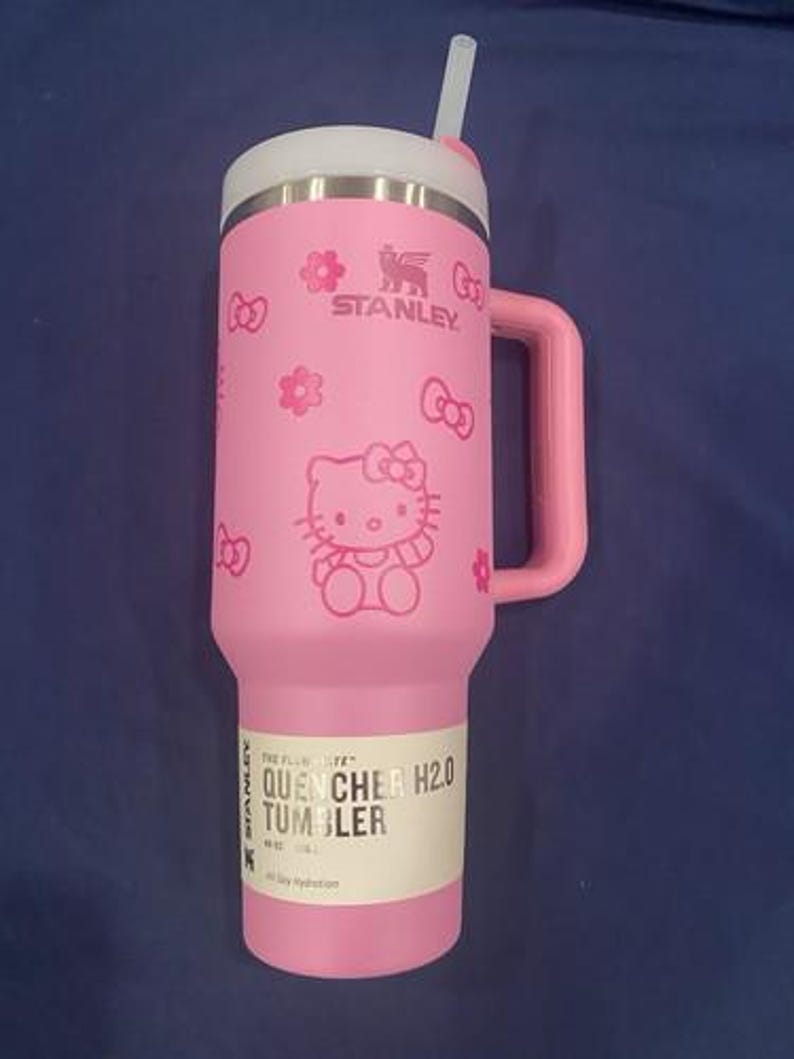 May include: A pink Stanley Quencher H2.0 tumbler featuring a Hello Kitty graphic with bows and flowers. The cup has a side handle, a white lid, and a clear straw. A product label on the lower section reads The FlowState Quencher H2.0 Tumbler 40 oz.