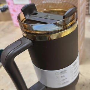 May include: A brown and white Stanley travel mug with a black handle and a clear lid. The mug has a gold-colored rim and a white band around the middle. The Stanley logo is visible on the packaging in the background.