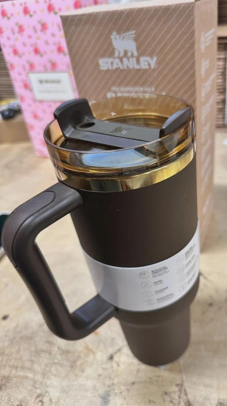 May include: A brown and white Stanley travel mug with a black handle and a clear lid. The mug has a gold-colored rim and a white band around the middle. The Stanley logo is visible on the packaging in the background.