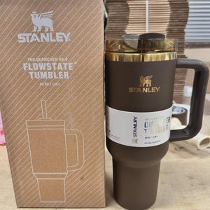 May include: A brown Stanley Flowstate tumbler with a handle and a gold-colored rim. The tumbler is next to its cardboard box, which features the Stanley logo and product details. The tumbler holds 40 oz.