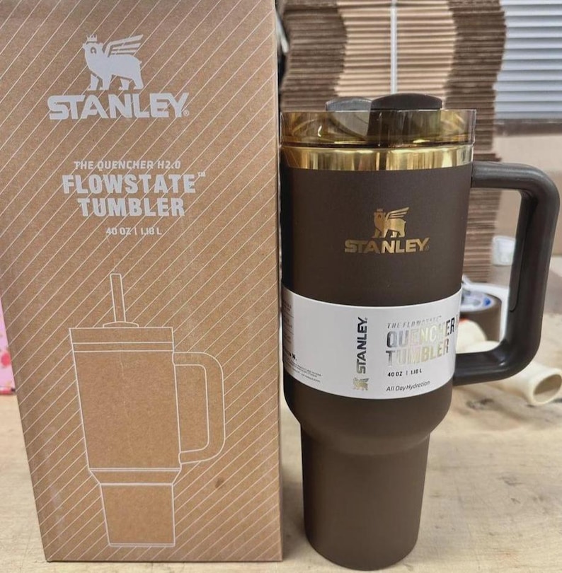 May include: A brown Stanley Flowstate tumbler with a handle and a gold-colored rim. The tumbler is next to its cardboard box, which features the Stanley logo and product details. The tumbler holds 40 oz.