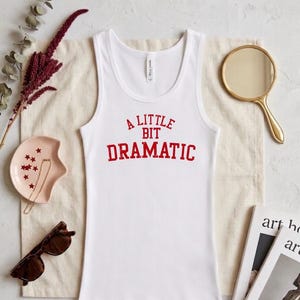 May include: A white sleeveless tank top featuring the red text A LITTLE BIT DRAMATIC printed in a bold, slightly distressed font across the chest. The garment has a scoop neckline and a standard fit, presented as a clean, simple piece of apparel.
