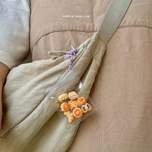 May include: A beige crossbody bag with a wide strap and a clear plastic pouch attached by a light purple clip. The pouch contains small, assorted orange and tan cookie-shaped charms. The text HAPPY AT HOME CLUB is printed on the bag fabric.