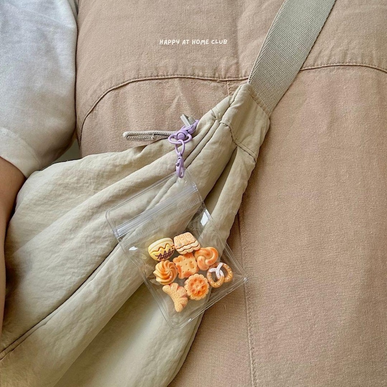 May include: A beige crossbody bag with a wide strap and a clear plastic pouch attached by a light purple clip. The pouch contains small, assorted orange and tan cookie-shaped charms. The text HAPPY AT HOME CLUB is printed on the bag fabric.