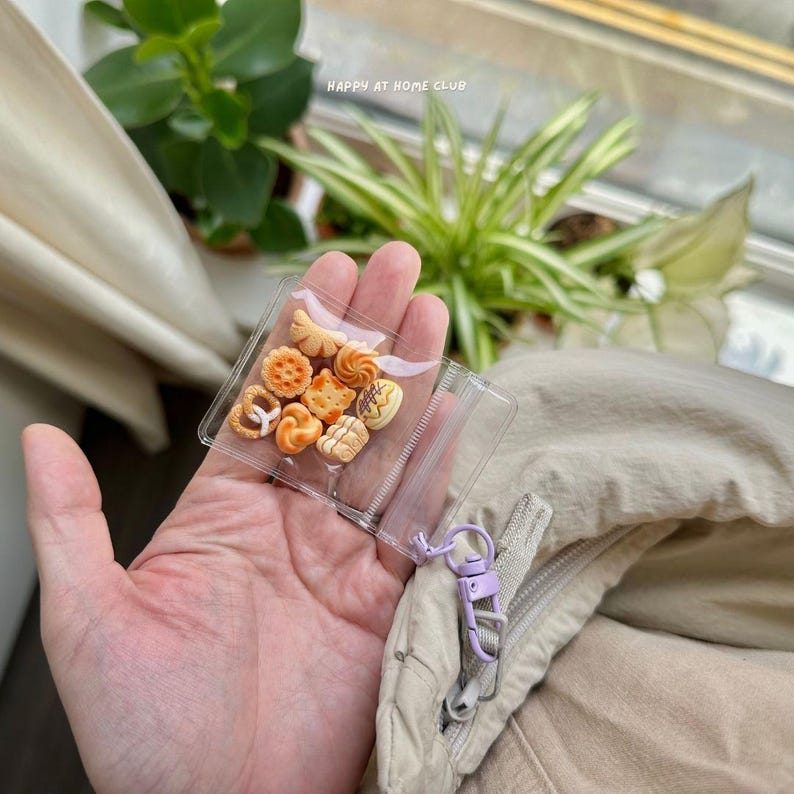 May include: A small, clear, square plastic pouch containing a collection of miniature, tan-coloured baked goods, including a pretzel, biscuits, and rolls. The pouch features a light purple metal lobster clasp attached to a zip pull for easy hanging.