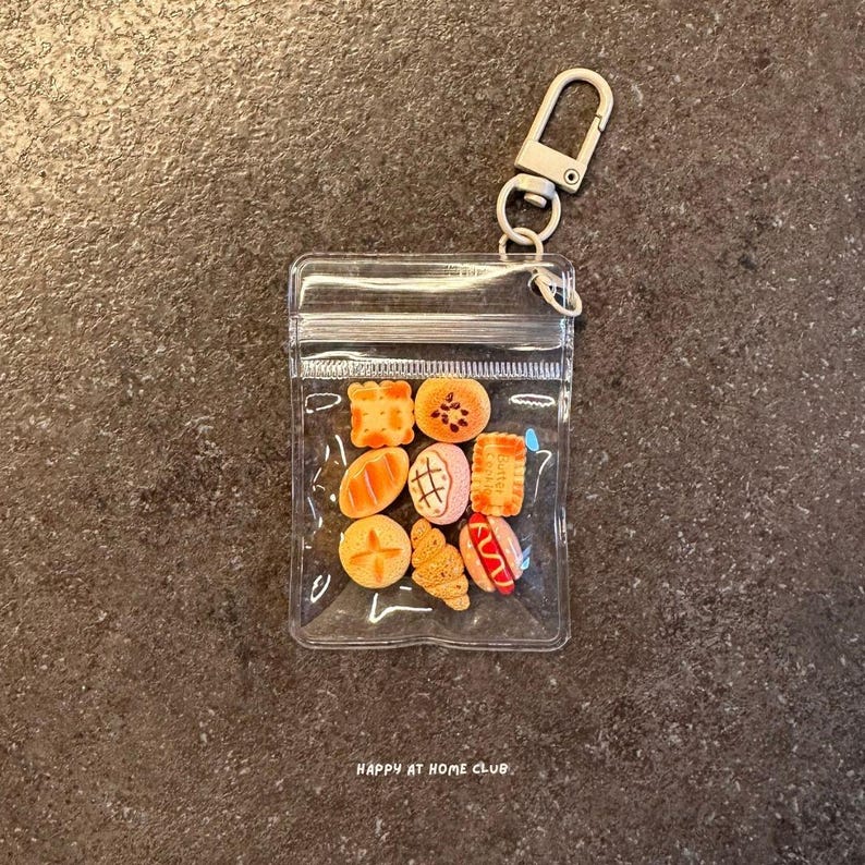 May include: A clear plastic zipped pouch keyring containing a collection of miniature food-shaped charms, including various breads, a hot dog, and biscuits. The metal hardware includes a lobster clasp and a ring for attachment to keys or bags.