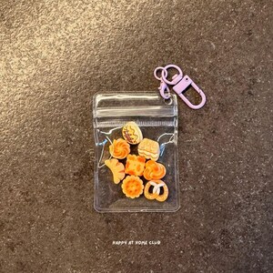 May include: A clear plastic pouch containing eight miniature orange and tan food-shaped charms, including a pretzel, cookie, and pastry shapes. The pouch features a light purple lobster clasp keychain attached to the top corner and the text HAPPY AT HOME CLUB.