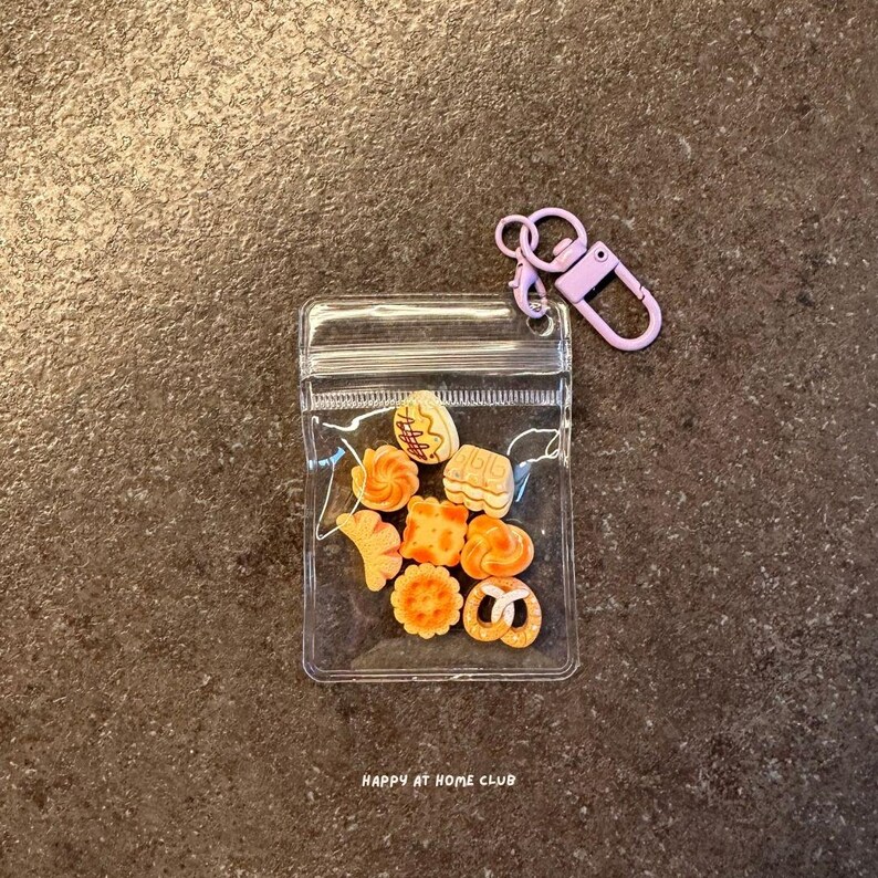 May include: A clear plastic pouch containing eight miniature orange and tan food-shaped charms, including a pretzel, cookie, and pastry shapes. The pouch features a light purple lobster clasp keychain attached to the top corner and the text HAPPY AT HOME CLUB.