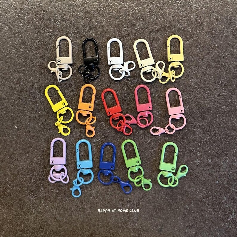 May include: A collection of fourteen metal swivel lobster claw clasps in various colours including black, white, yellow, orange, red, pink, purple, blue, and green, each featuring a small attached split ring, with the text HAPPY AT HOME CLUB.